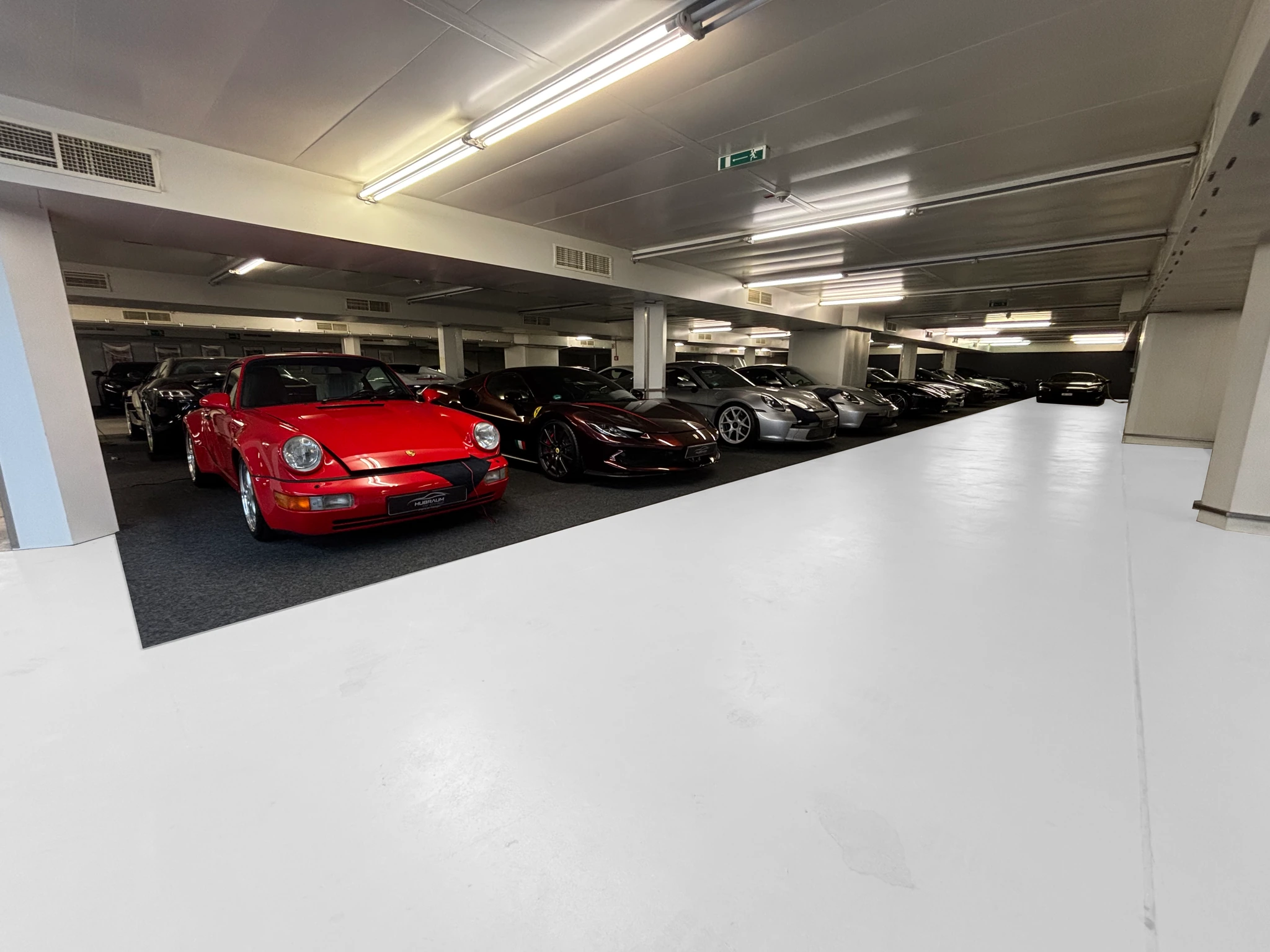 Stellplatzreihe unter Leuchtstoffröhren in der HUBRAUM-Halle: ein roter Porsche 911, ein dunkler Ferrari und weitere Fahrzeuge, davor die helle Fahrfläche