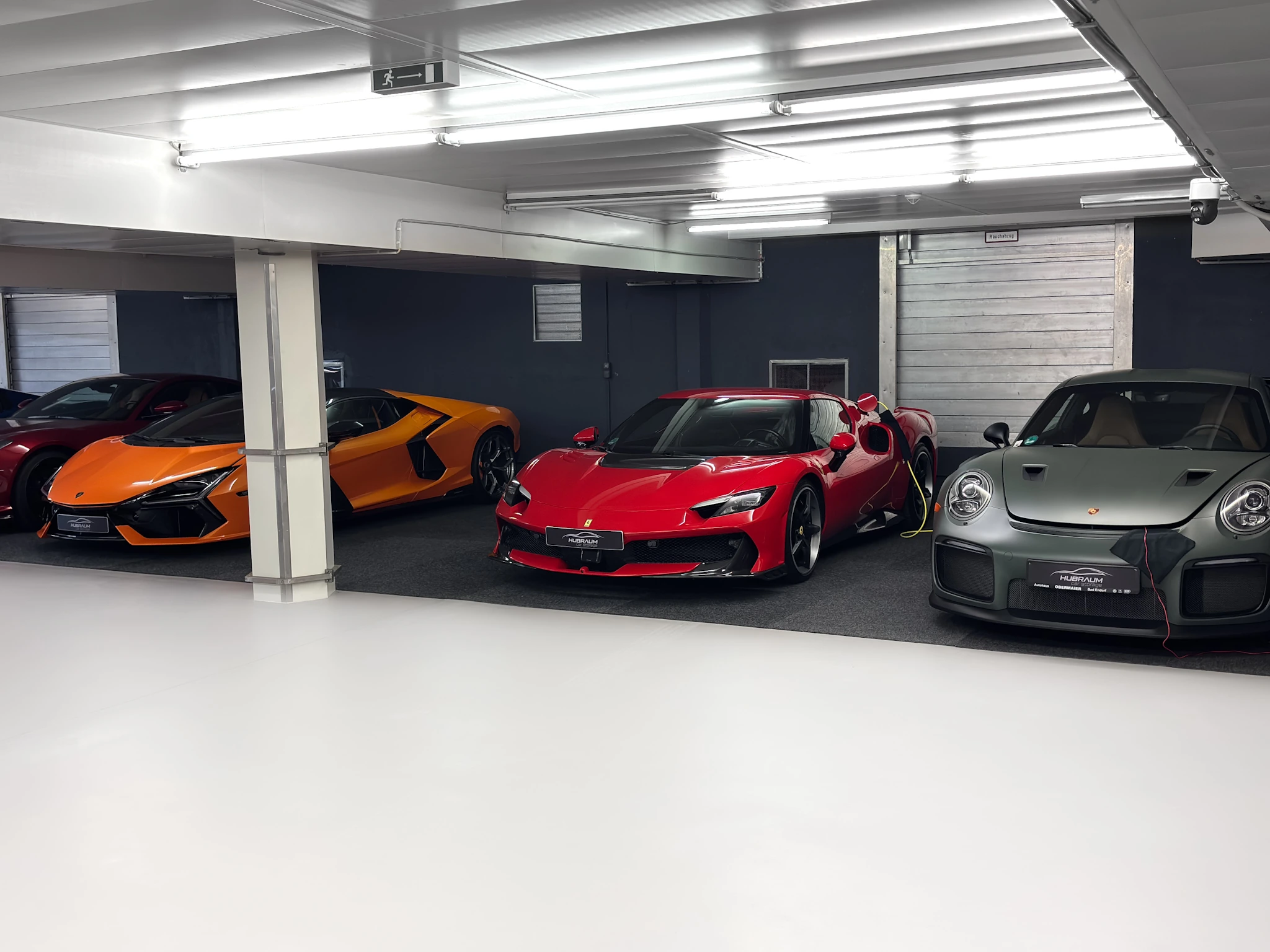 Drei Stellplätze in der HUBRAUM-Halle: ein orangefarbener Lamborghini, ein roter Ferrari und ein grüner Porsche 911, jedes Fahrzeug mit angeschlossenem Erhaltungsladegerät