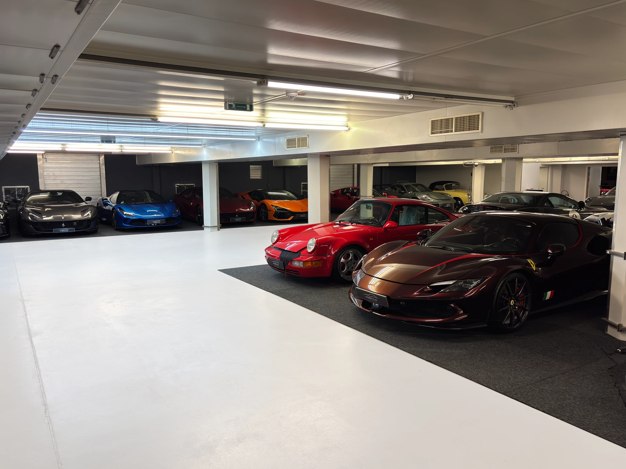 Weite Sicht in die HUBRAUM-Halle: vorn ein dunkelroter Ferrari und ein roter Porsche 911, dahinter Stellplätze mit einem blauen Ferrari und einem orangefarbenen McLaren