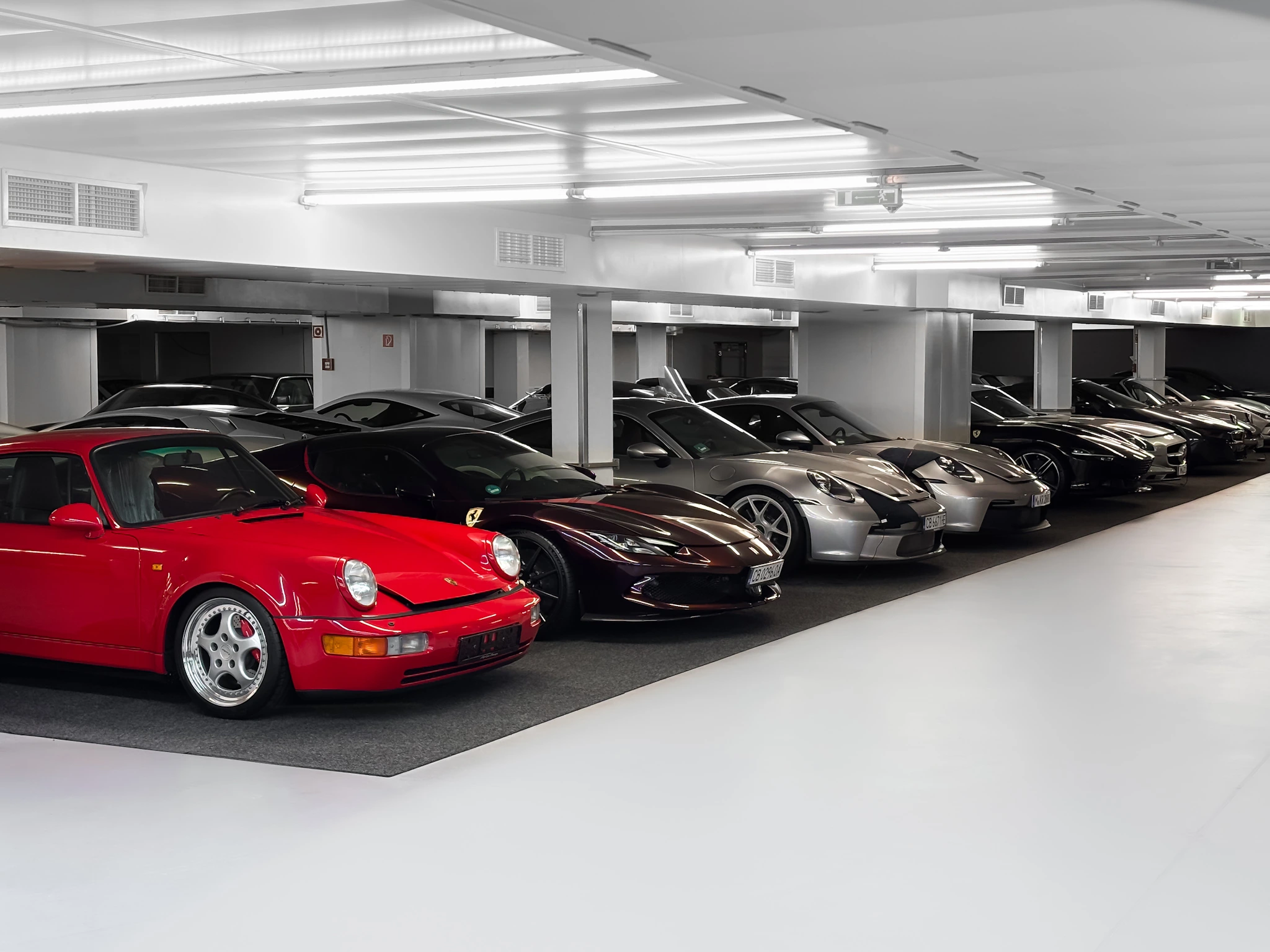 Stellplatzreihe in der HUBRAUM-Halle: vorn ein roter Porsche 911, dahinter ein dunkelroter Ferrari, ein silberner Porsche und weitere Sportwagen, davor die helle, versiegelte Fahrfläche